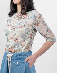 A woman wearing a beige short-sleeve top with a blue and pink floral paisley pattern, paired with blue denim shorts, against a plain light gray background.