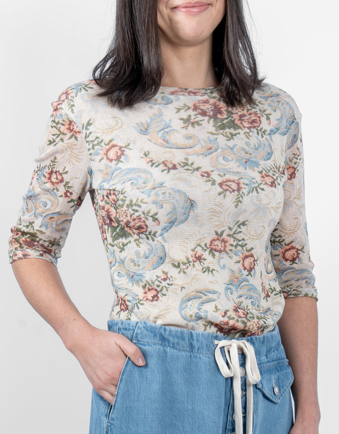 A woman wearing a three-quarter sleeve top with a vintage floral and scroll pattern in beige, blue, and pink, paired with light blue denim shorts against a plain white background.