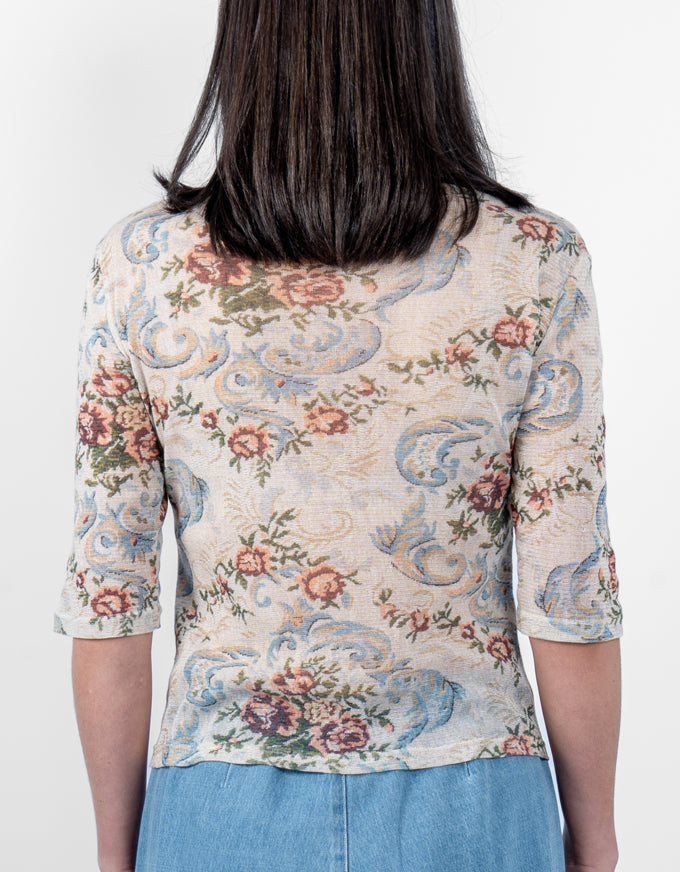 A woman viewed from the back wearing a short-sleeve top with a beige base and a detailed floral and scroll pattern in blue, pink, and green, paired with light blue jeans against a plain white background.