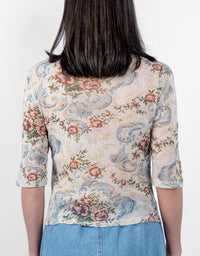 A woman viewed from the back wearing a short-sleeve top with a beige base and a detailed floral and scroll pattern in blue, pink, and green, paired with light blue jeans against a plain white background.