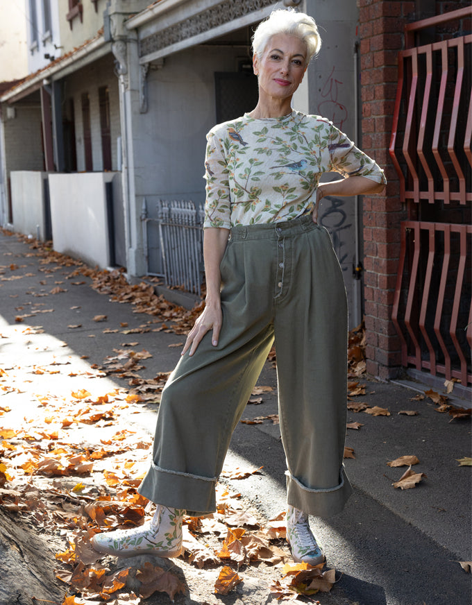 A woman stands on an outdoor sidewalk in a city street, wearing a light beige floral long-sleeve top and high-waisted olive green wide-leg trousers with rolled cuffs, paired with white sneakers.