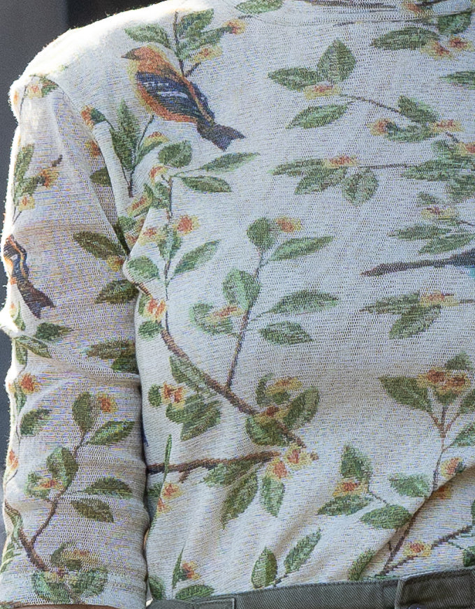 Close-up of a woven textile featuring a botanical pattern with green leaves, brown branches, and small birds in blue, orange, and yellow on an off-white background.