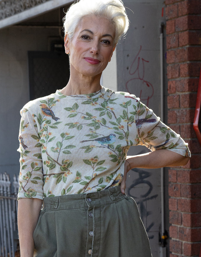 A woman with short platinum blonde hair wearing a beige three-quarter sleeve top with a green and blue bird and leaf pattern, paired with olive green high-waisted trousers, standing outdoors against a brick and concrete wall.