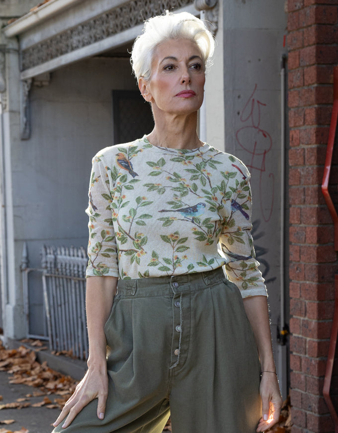 A woman with short white hair stands outdoors wearing a cream-colored long-sleeve top with a green and blue bird and leaf pattern, paired with olive green high-waisted trousers.