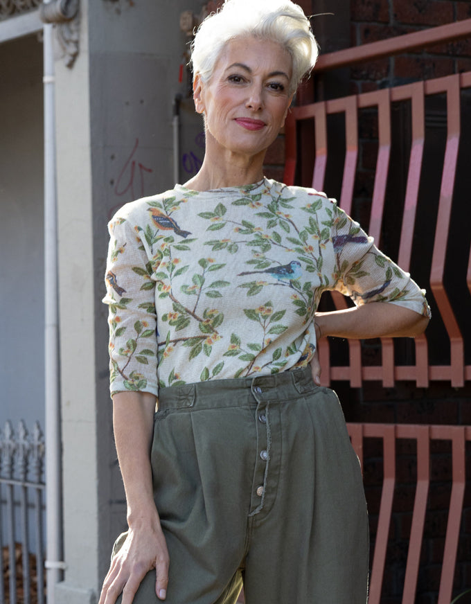 A woman with short white hair stands outdoors wearing a cream-colored short-sleeve blouse with a green and blue bird and leaf print, paired with olive green high-waisted trousers.