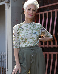 A woman with short white hair stands outdoors wearing a cream-colored short-sleeve blouse with a green and blue bird and leaf print, paired with olive green high-waisted trousers.