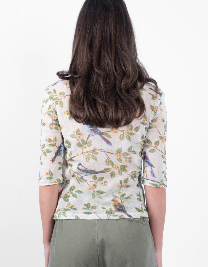 A woman viewed from behind wearing a cream-colored three-quarter sleeve top with a detailed bird and leaf print, paired with olive green trousers, against a plain white background.