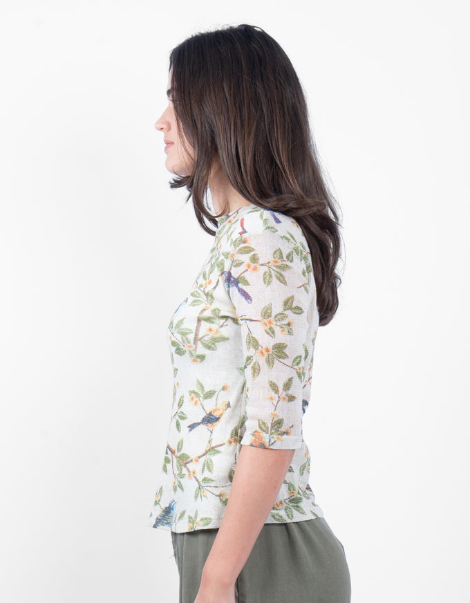 A woman shown in profile wearing a short-sleeve top with a white base and a colorful bird and leaf print, paired with olive green pants, against a plain white background.