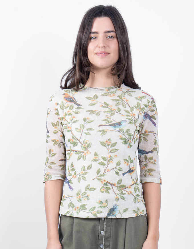 A woman modeling a three-quarter sleeve top with a delicate bird and leaf pattern on a light cream background, paired with olive green pants, against a plain white studio background.