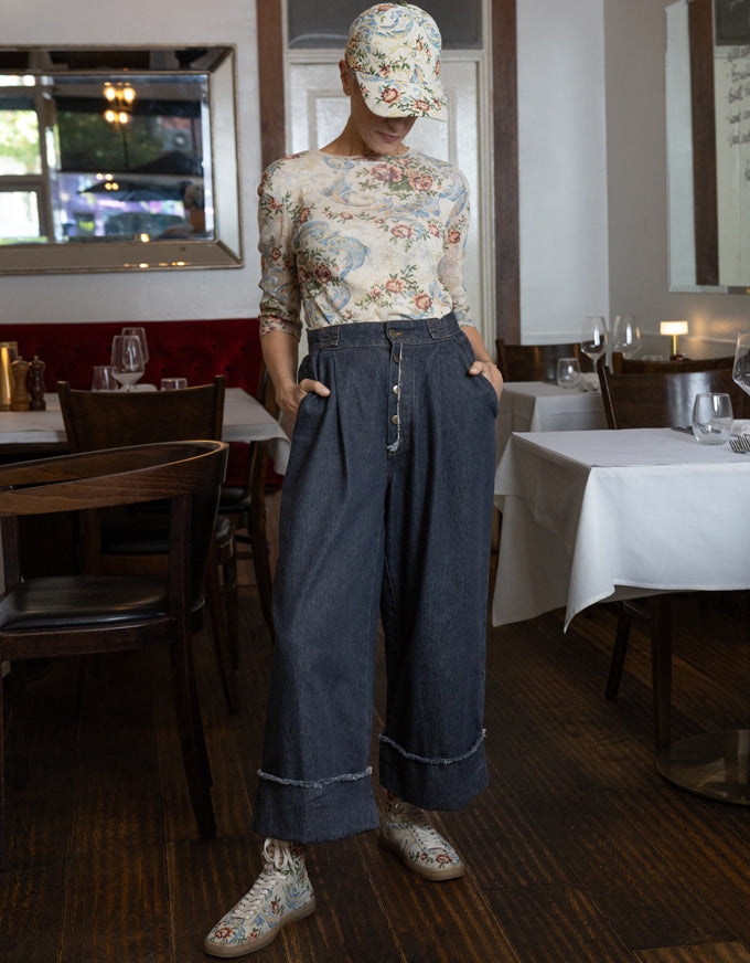 A person standing in a restaurant wearing a beige floral long-sleeve top, dark wide-leg denim pants with rolled cuffs, a matching beige floral cap, and beige floral lace-up shoes.