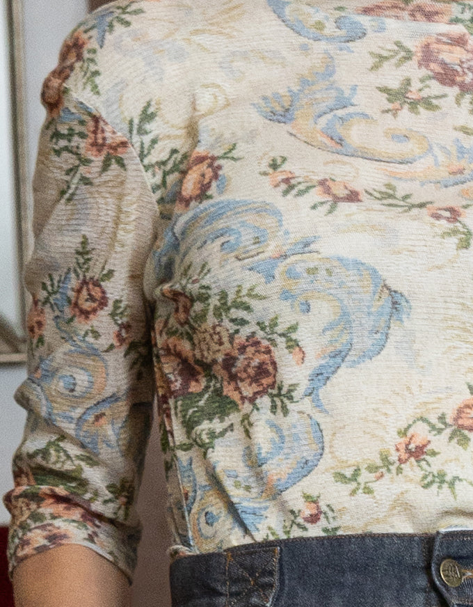 A close-up of a person wearing a textured, vintage-style floral and scroll pattern top with blue, green, and pink details on a cream background, paired with dark denim jeans.