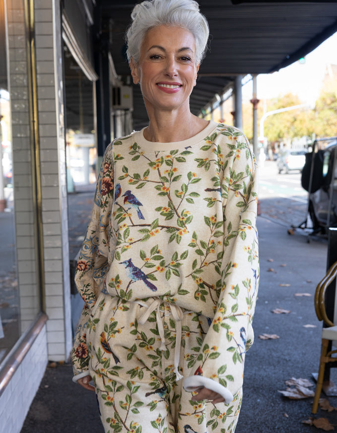 A smiling woman with short gray hair stands on a city sidewalk wearing a cream-colored two-piece lounge set with a green leafy branch pattern and blue birds. The set includes a long-sleeve top and matching drawstring pants.