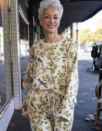 A smiling woman with short gray hair stands on a city sidewalk wearing a cream-colored two-piece lounge set with a green leafy branch pattern and blue birds. The set includes a long-sleeve top and matching drawstring pants.