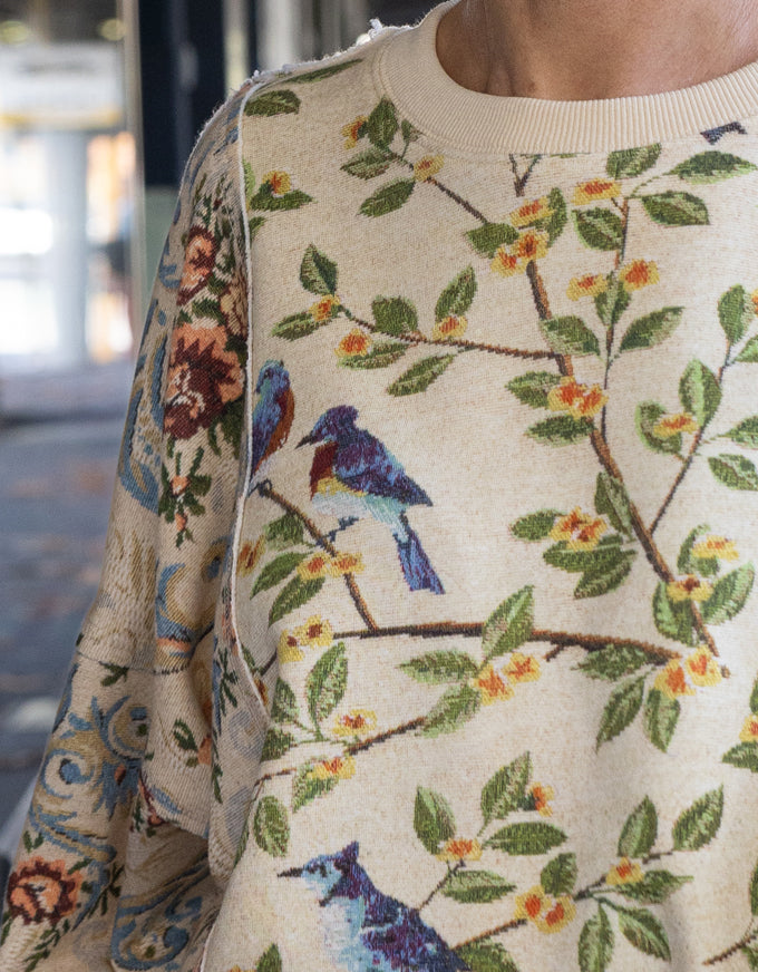 A close-up of a cream-colored knit sweater featuring a detailed botanical and avian pattern with green leaves, yellow-orange blossoms, and blue birds, worn by a model.