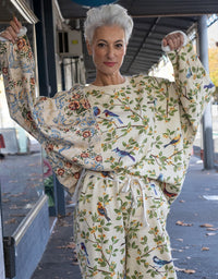 An older woman with short white hair stands outdoors under a covered walkway, wearing a cream-colored two-piece pajama set with a detailed print of blue birds, green leaves, and orange flowers. She holds up both sleeves of the matching long-sleeve top to show the fit and pattern.