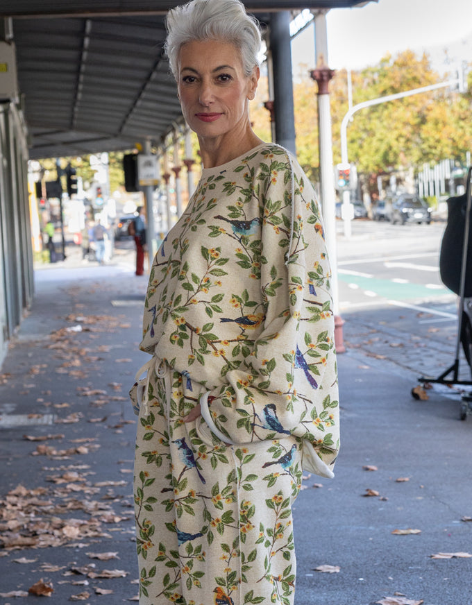 A woman with short gray hair stands on a city sidewalk wearing a cream-colored robe or kimono-style outfit with an all-over print of green leaves, orange flowers, and blue birds.