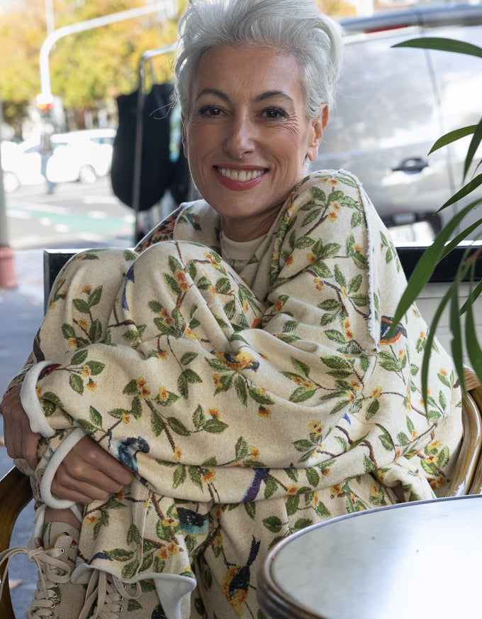 A smiling woman with short gray hair sits wrapped in a cream-colored throw blanket featuring a green leafy vine pattern and small orange and blue bird motifs. The setting appears to be a cafe or outdoor seating area with a blurred street background.