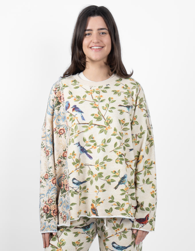 A woman modeling a matching pajama set with a cream-colored base and a detailed botanical and bird pattern, featuring bluebirds, leaves, and floral tapestry-style panels.