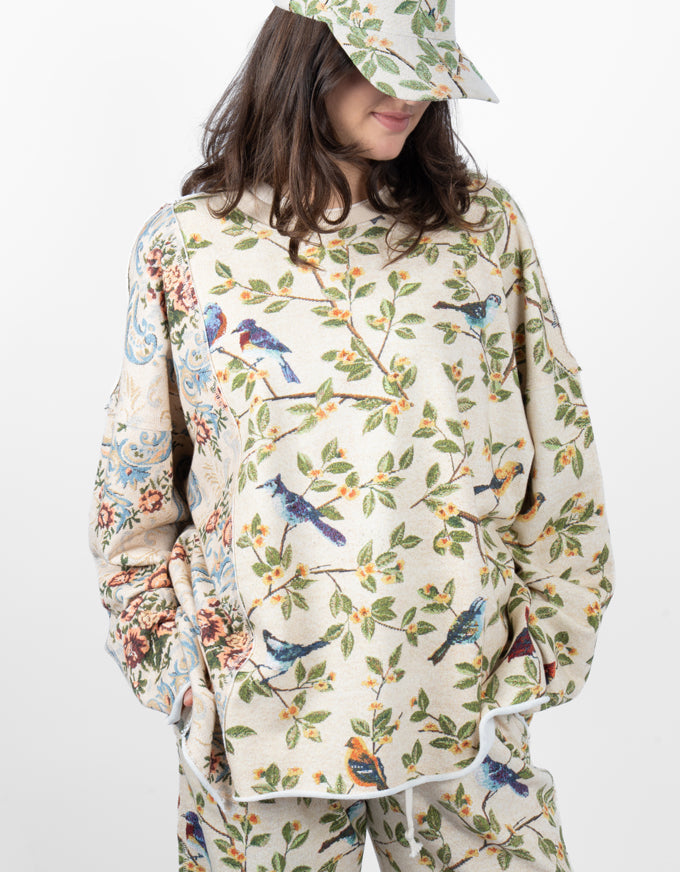 A woman wearing a cream-colored long-sleeve top and matching pants with a detailed bird and floral print, paired with a matching cap, against a plain white background.