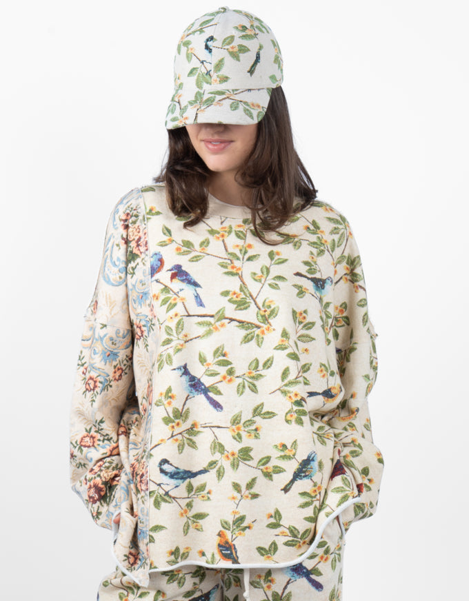 A model wearing a matching two-piece pajama set and a baseball cap, featuring a cream-colored base with a detailed print of blue birds, green leaves, and orange berries.