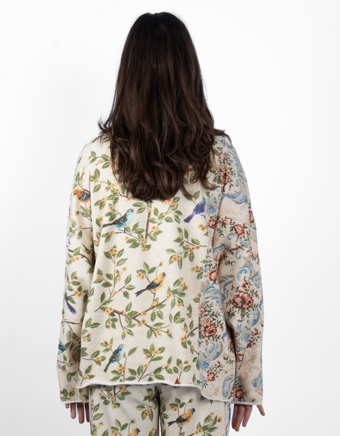 A woman viewed from behind wearing a matching pajama set with a split design: one side features a cream background with green leaves, blue birds, and small yellow flowers, while the other side has a blue and pink floral pattern. She is standing against a plain white background.