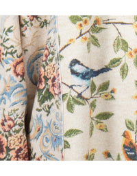 Close-up of a woven textile with a floral and bird pattern on a light beige background, showing branches with green leaves, small yellow flowers, and blue-and-black birds.