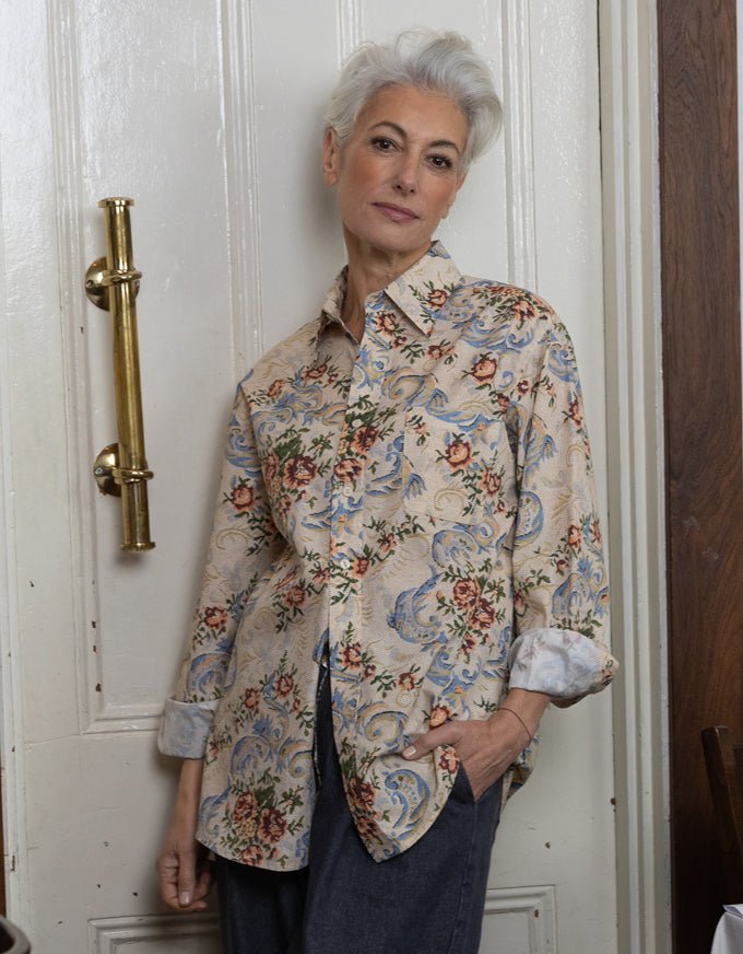 An older woman with short gray hair stands indoors wearing a beige button-up shirt with a blue, red, and green floral and scroll pattern, paired with dark blue jeans. She is posed in front of a white paneled door with a brass handle.