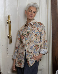 An older woman with short gray hair stands indoors wearing a beige button-up shirt with a blue, red, and green floral and scroll pattern, paired with dark blue jeans. She is posed in front of a white paneled door with a brass handle.