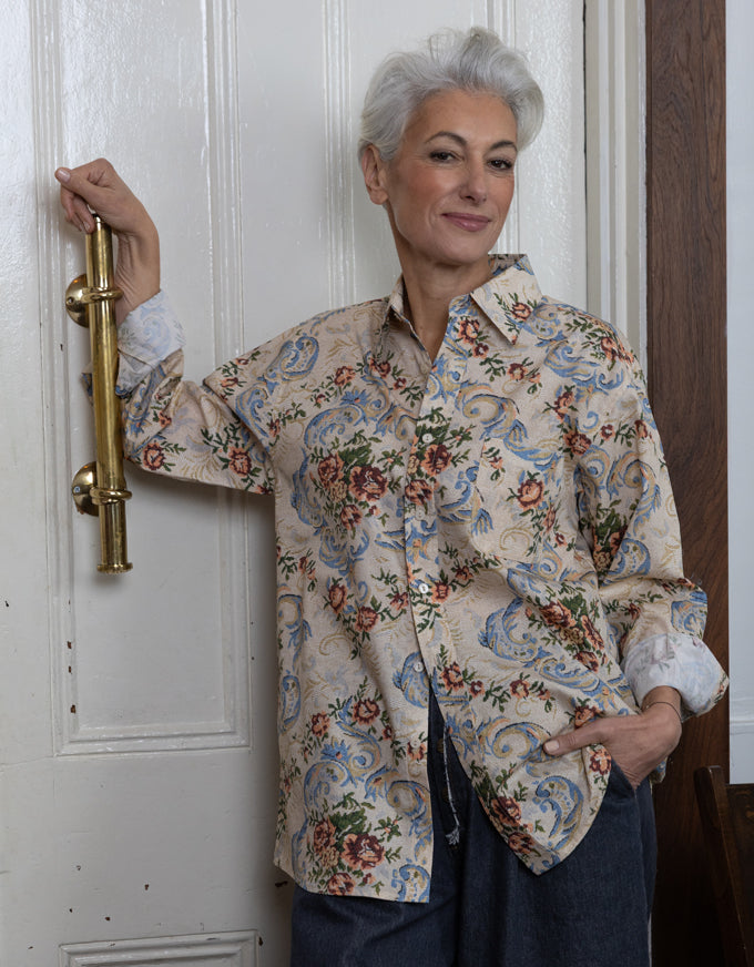 A woman with short gray hair stands in front of a white paneled door, wearing a long-sleeve button-up shirt with a beige base and a blue, red, and green floral and paisley pattern, paired with dark blue pants.