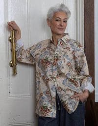 A woman with short gray hair stands in front of a white paneled door, wearing a long-sleeve button-up shirt with a beige base and a blue, red, and green floral and paisley pattern, paired with dark blue pants.