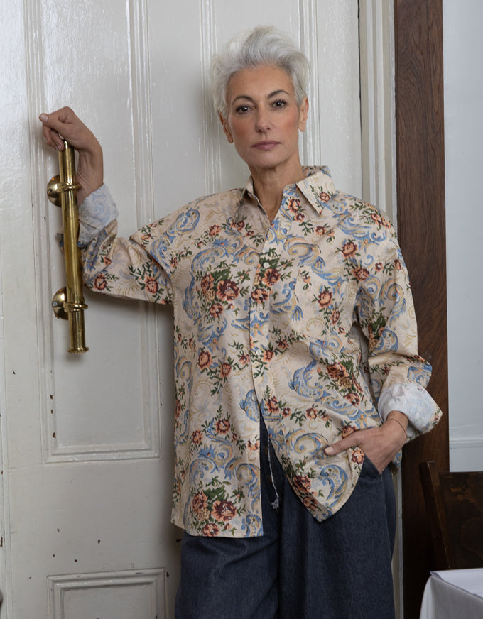 A woman with short white hair stands indoors against a white paneled door, wearing a long-sleeve button-up shirt with a beige base and an ornate multicolor floral and paisley print, paired with dark blue pants.