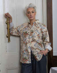 A woman with short white hair stands indoors against a white paneled door, wearing a long-sleeve button-up shirt with a beige base and an ornate multicolor floral and paisley print, paired with dark blue pants.