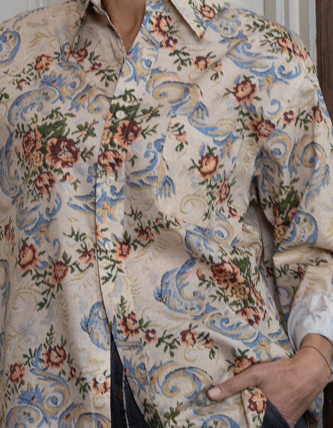 A close-up of a person wearing a long-sleeve button-up shirt with a dense floral and scroll pattern in blue, green, and rust-brown on a light beige background.