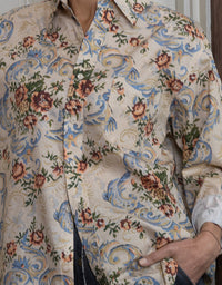 A close-up of a person wearing a long-sleeve button-up shirt with a dense floral and scroll pattern in blue, green, and rust-brown on a light beige background.