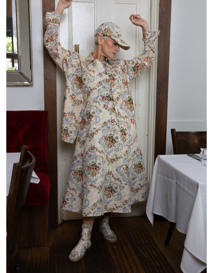 A woman wearing a long-sleeved cream-colored dress with a colorful floral and paisley pattern, a matching cap, and matching socks, posing in a doorway of a restaurant or cafe.