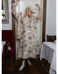 A woman wearing a long-sleeved cream-colored dress with a colorful floral and paisley pattern, a matching cap, and matching socks, posing in a doorway of a restaurant or cafe.