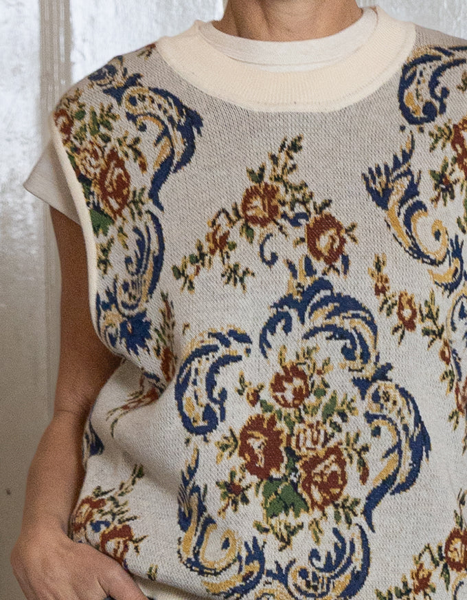 A close-up of a person wearing a cream knit sweater vest with a blue, brown, and green floral damask pattern over a white crewneck shirt.
