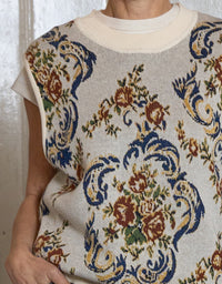 A close-up of a person wearing a cream knit sweater vest with a blue, brown, and green floral damask pattern over a white crewneck shirt.