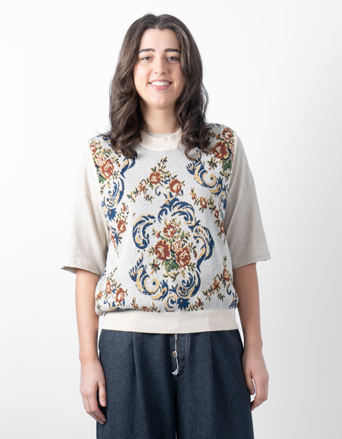 A woman modeling a short-sleeve knit top with a cream base and a large, colorful floral and scroll pattern in blue, rust orange, and green, paired with dark blue wide-leg pants against a plain white background.