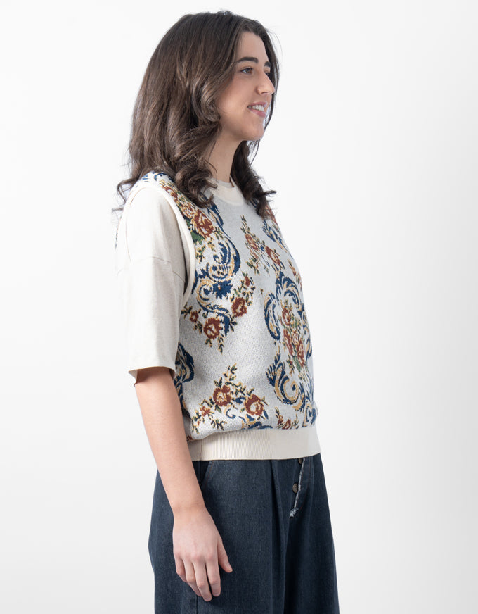A woman shown in profile wearing a cream short-sleeve top layered under a sleeveless knit vest with a multicolor floral and scroll pattern, paired with dark wide-leg pants, against a plain white studio background.