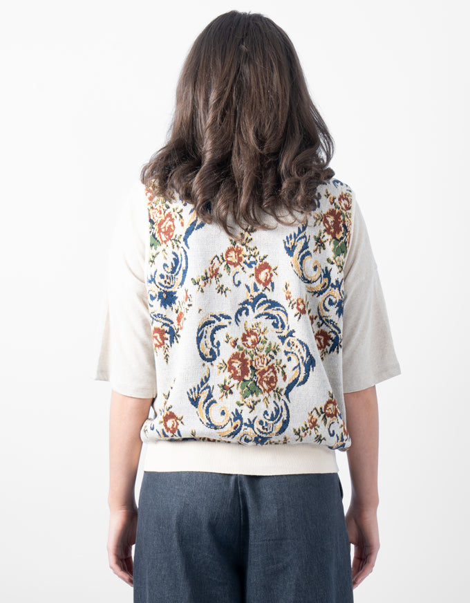 A woman shown from behind wearing a cream-colored short-sleeve top with a highly detailed, colorful floral and scrollwork tapestry-style pattern across the back, paired with dark blue jeans.