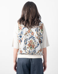A woman shown from behind wearing a cream-colored short-sleeve top with a highly detailed, colorful floral and scrollwork tapestry-style pattern across the back, paired with dark blue jeans.