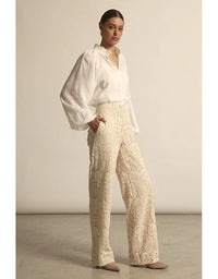 Woman wearing a white blouse and beige lace pants against a plain background