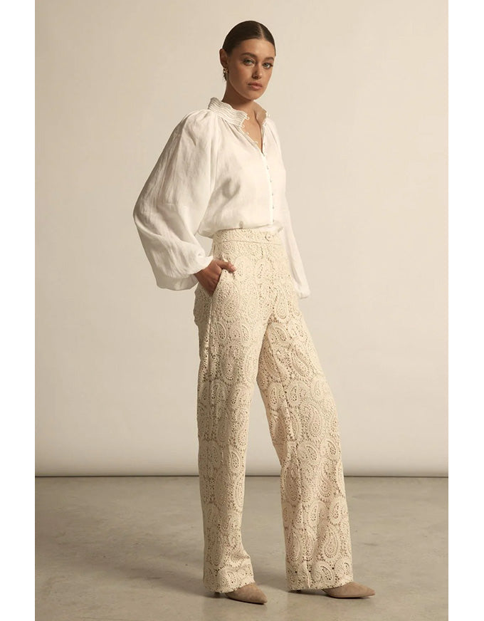 Woman wearing a white blouse and beige lace pants against a plain background