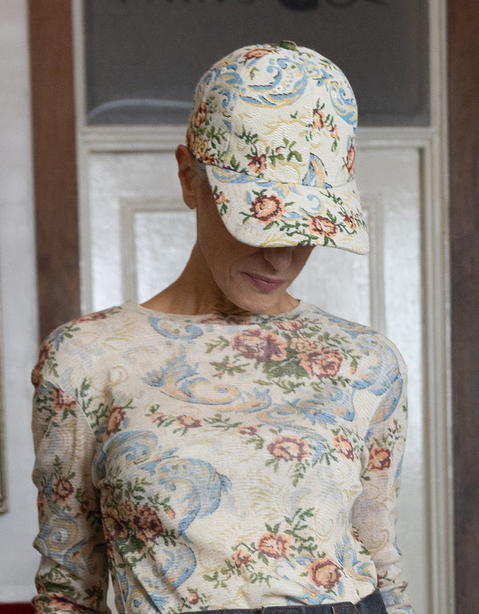 A woman wearing a matching beige floral cap and long-sleeve top, looking down. The fabric has a detailed pattern of blue scrolls, pink roses, and green leaves on a cream background.