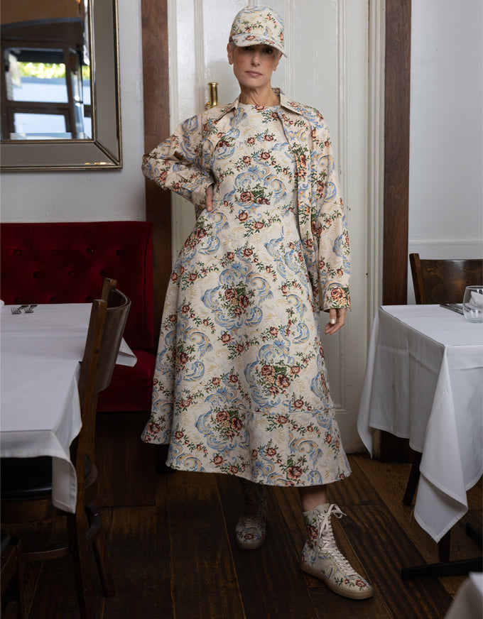 A model stands in a restaurant wearing a long-sleeved midi dress with a beige, blue, and red floral pattern, a matching cap, and lace-up boots.