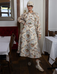 A model stands in a restaurant wearing a long-sleeved midi dress with a beige, blue, and red floral pattern, a matching cap, and lace-up boots.