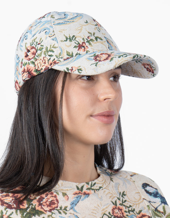 A woman wearing a beige woven baseball cap with a floral and bird pattern, shown in a close-up profile shot against a plain white background.