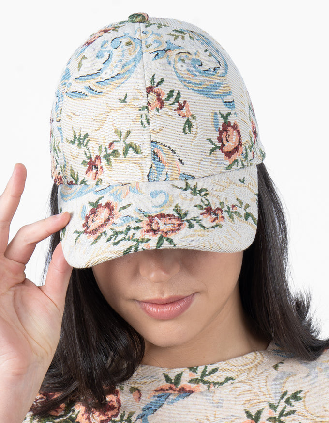 A woman wearing a structured baseball cap with a detailed floral and scroll pattern in muted blue, pink, and green on a cream background. She is holding the brim with her hand against a clean white background.