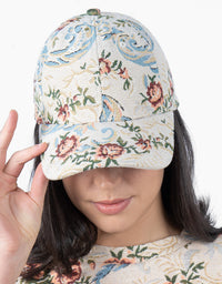 A woman wearing a structured baseball cap with a detailed floral and scroll pattern in muted blue, pink, and green on a cream background. She is holding the brim with her hand against a clean white background.